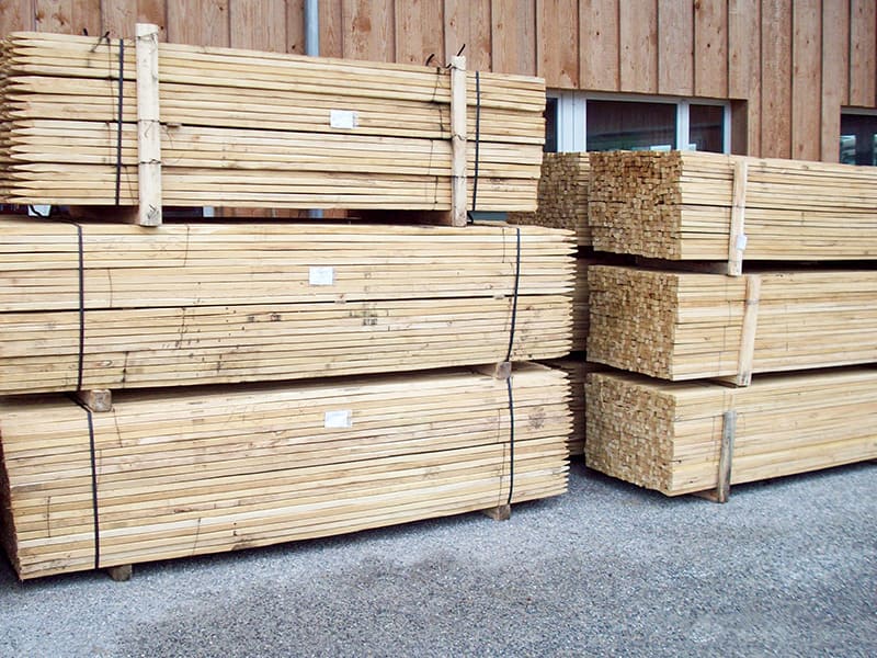 Gestapelte, gebündelte Robinienholzpfähle vor einer Holzfassade auf Asphaltboden.